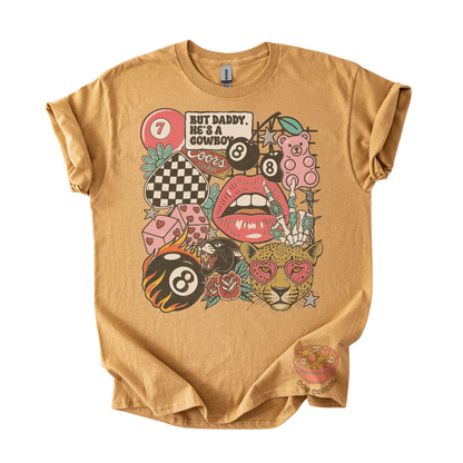 A tan graphic t-shirt featuring a colorful retro-style print with billiards, lips, a leopard, and the text 'BUT DADDY, HE'S A COWBOY'.