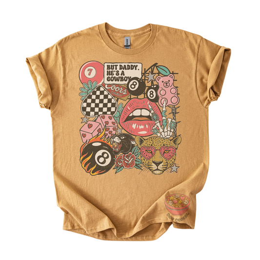 A tan graphic t-shirt featuring a colorful retro-style print with billiards, lips, a leopard, and the text 'BUT DADDY, HE'S A COWBOY'.
