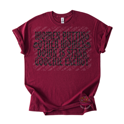 A maroon short-sleeve graphic t-shirt laid flat on a white background. The shirt features a distressed black text graphic reading 'WOMEN PUTTING OTHER WOMEN DOWN IS STAY COCHCHI ENERGY' across the chest.