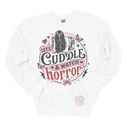 White crewneck sweatshirt featuring a spooky cute horror-inspired design with the phrase "Let's Cuddle & Watch Horror," pink hearts, butterflies, bows, and a creepy long-haired figure emerging from a television.