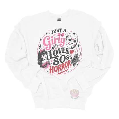 White crewneck sweatshirt featuring the phrase "Just a Girly Who Loves 80s Horror" with retro horror-inspired artwork including a hockey mask, clawed glove, vintage girl illustration, pink bows, and spooky accents.
