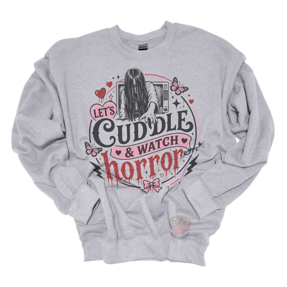 Light grey crewneck sweatshirt featuring a spooky cute horror-inspired design with the phrase "Let's Cuddle & Watch Horror," pink hearts, butterflies, bows, and a creepy long-haired figure emerging from a television.