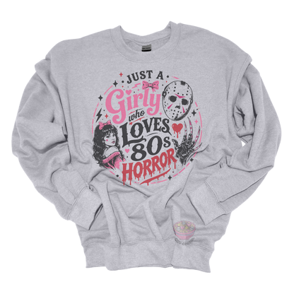Heather gray crewneck sweatshirt featuring the phrase "Just a Girly Who Loves 80s Horror" with retro horror-inspired artwork including a hockey mask, clawed glove, vintage girl illustration, pink bows, and spooky accents.