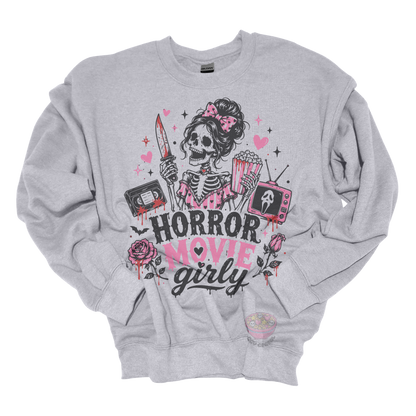 Gray crewneck sweatshirt featuring a skeleton with a pink bow holding a bloody knife and popcorn, surrounded by horror-inspired movie details, roses, hearts, a VHS tape, retro television, and the phrase "Horror Movie Girly."