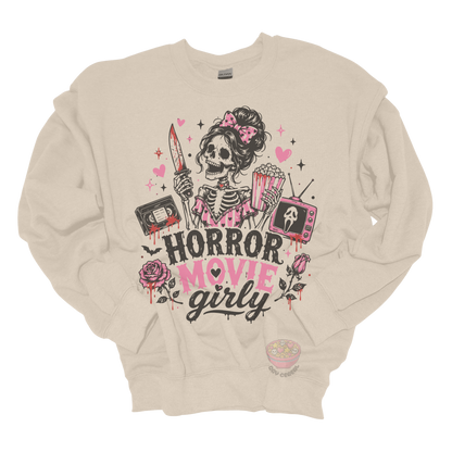Beige crewneck sweatshirt featuring a skeleton with a pink bow holding a bloody knife and popcorn, surrounded by horror-inspired movie details, roses, hearts, a VHS tape, retro television, and the phrase "Horror Movie Girly."
