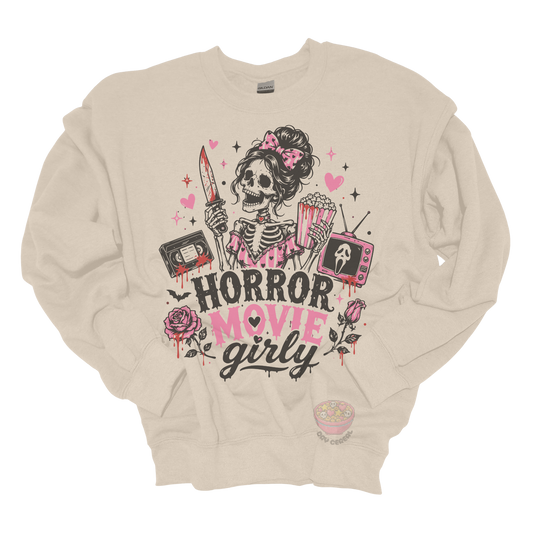 Beige crewneck sweatshirt featuring a skeleton with a pink bow holding a bloody knife and popcorn, surrounded by horror-inspired movie details, roses, hearts, a VHS tape, retro television, and the phrase "Horror Movie Girly."