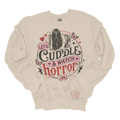 Beige crewneck sweatshirt featuring a spooky cute horror-inspired design with the phrase "Let's Cuddle & Watch Horror," pink hearts, butterflies, bows, and a creepy long-haired figure emerging from a television.
