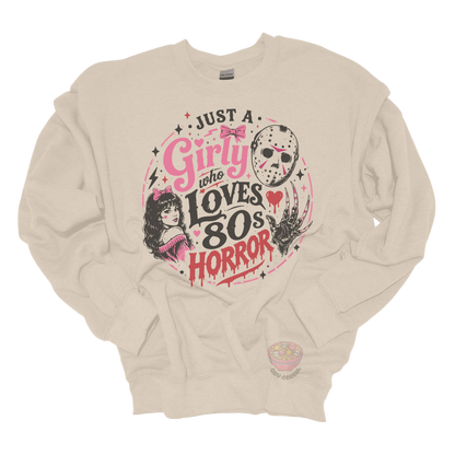 Beige crewneck sweatshirt featuring the phrase "Just a Girly Who Loves 80s Horror" with retro horror-inspired artwork including a hockey mask, clawed glove, vintage girl illustration, pink bows, and spooky accents.