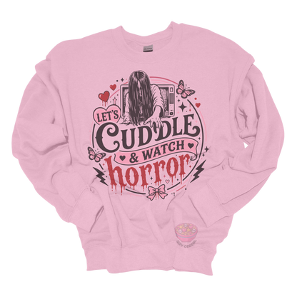 Light pink crewneck sweatshirt featuring a spooky cute horror-inspired design with the phrase "Let's Cuddle & Watch Horror," pink hearts, butterflies, bows, and a creepy long-haired figure emerging from a television.