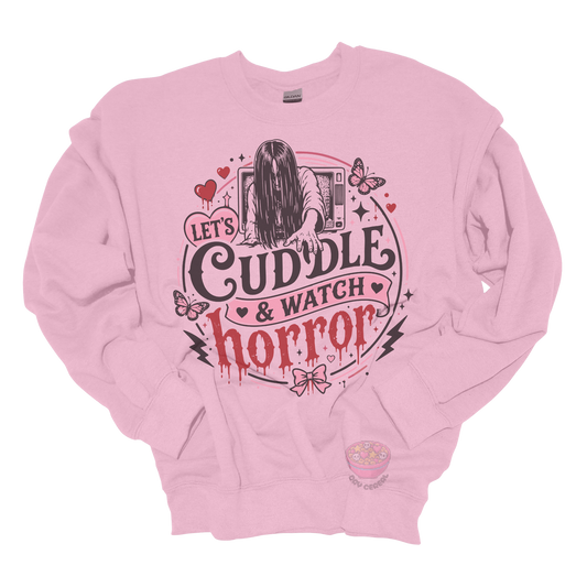 Light pink crewneck sweatshirt featuring a spooky cute horror-inspired design with the phrase "Let's Cuddle & Watch Horror," pink hearts, butterflies, bows, and a creepy long-haired figure emerging from a television.