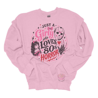 Light pink crewneck sweatshirt featuring the phrase "Just a Girly Who Loves 80s Horror" with retro horror-inspired artwork including a hockey mask, clawed glove, vintage girl illustration, pink bows, and spooky accents.