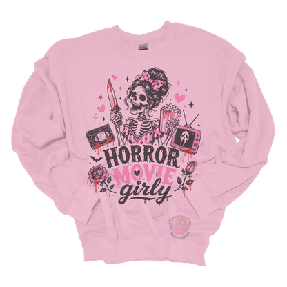 Light pink crewneck sweatshirt featuring a skeleton with a pink bow holding a bloody knife and popcorn, surrounded by horror-inspired movie details, roses, hearts, a VHS tape, retro television, and the phrase "Horror Movie Girly."