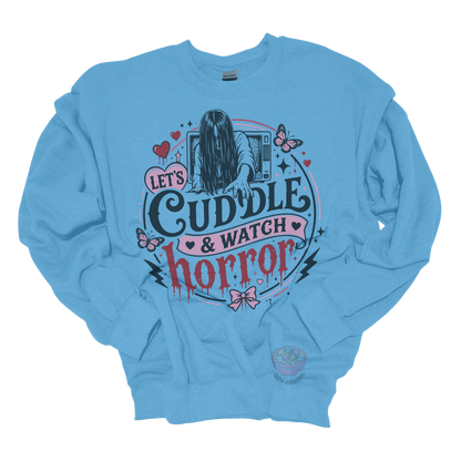 Light blue crewneck sweatshirt featuring a spooky cute horror-inspired design with the phrase "Let's Cuddle & Watch Horror," pink hearts, butterflies, bows, and a creepy long-haired figure emerging from a television.