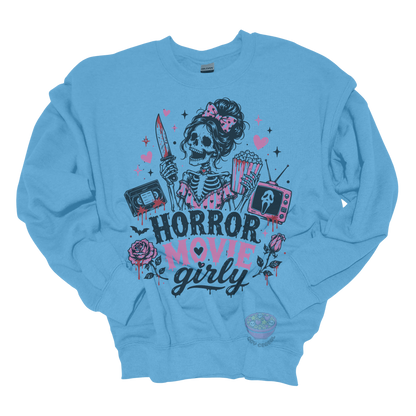 Light blue crewneck sweatshirt featuring a skeleton with a pink bow holding a bloody knife and popcorn, surrounded by horror-inspired movie details, roses, hearts, a VHS tape, retro television, and the phrase "Horror Movie Girly."