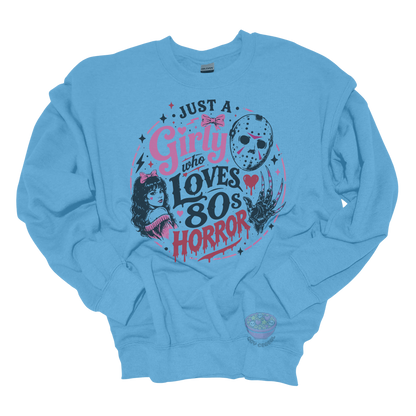 Light blue crewneck sweatshirt featuring the phrase "Just a Girly Who Loves 80s Horror" with retro horror-inspired artwork including a hockey mask, clawed glove, vintage girl illustration, pink bows, and spooky accents.
